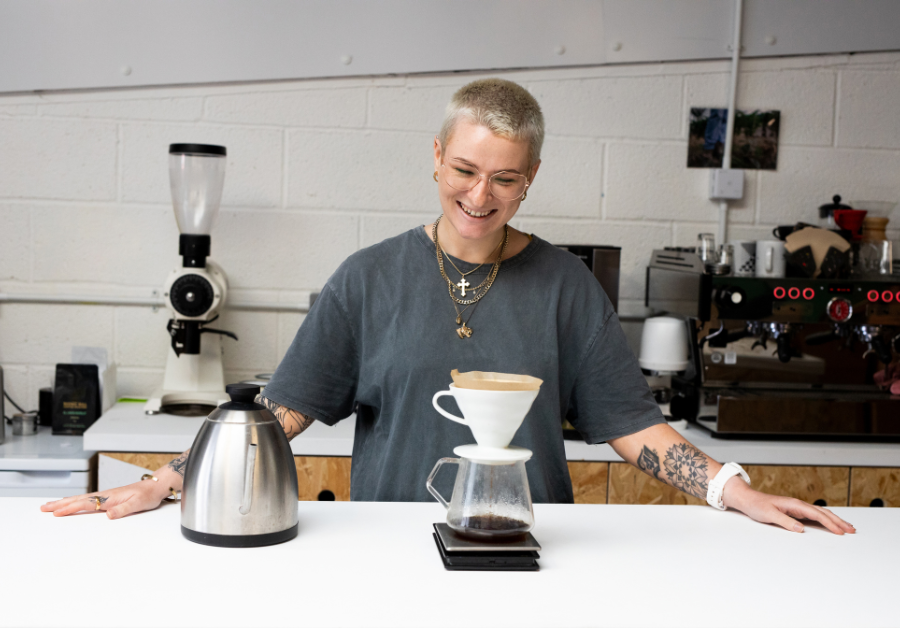 Hymyilevä henkilö valmistaa käsinsuodatettua kahvia pour over -menetelmällä kahvilan tiskin ääressä, vieressään vedenkeitin ja taustalla kahvikoneita.