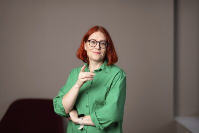 Annika Rönni-Sällinen seisoo neutraalin taustan edessä, katsoo kameraan hymyillen, toinen käsi on ristissä ja toinen osoittaa eteenpäin.