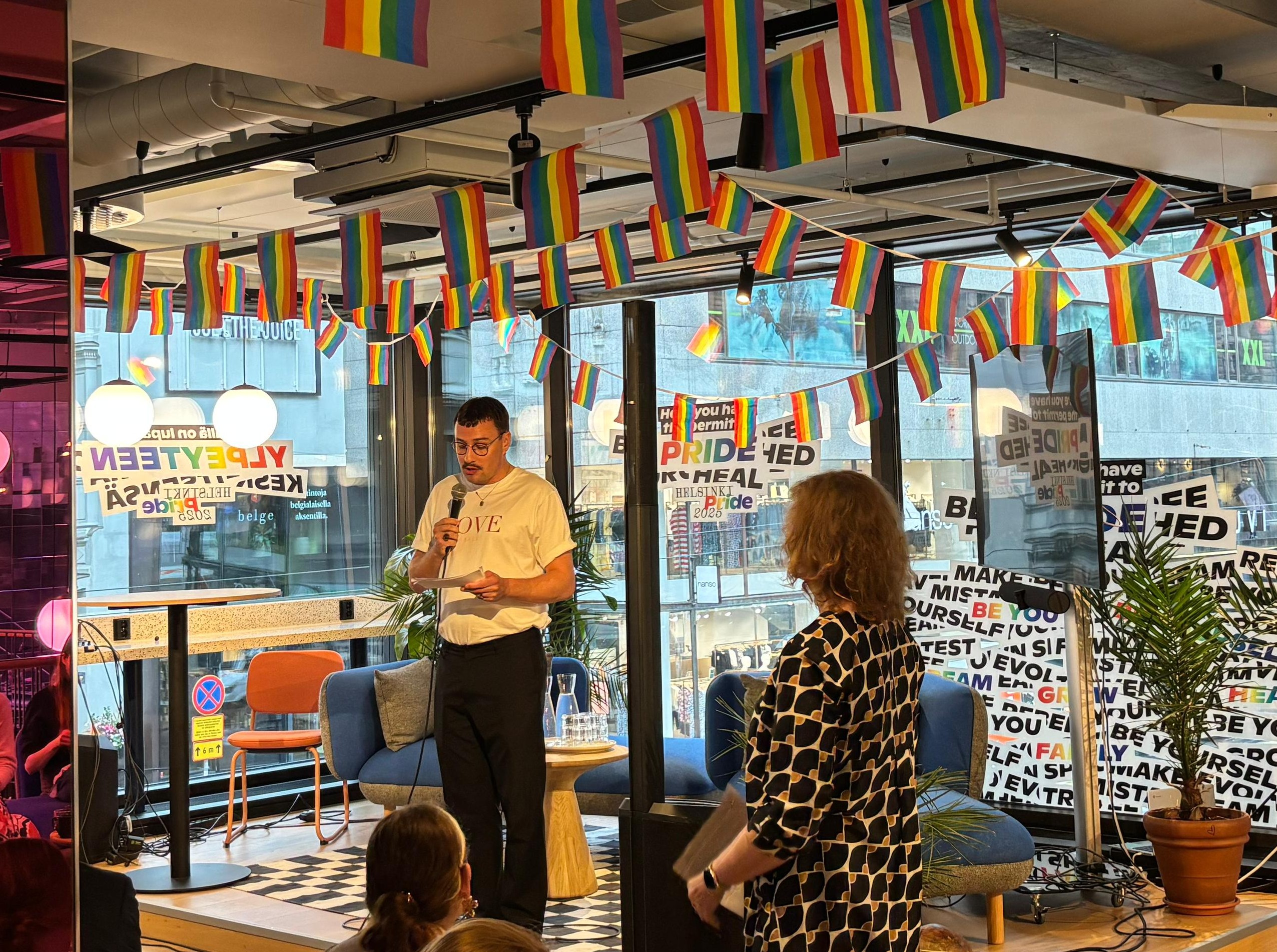 Kuvan keskellä juontaja Joonas Pesonen seisoo lavalla ja puhuu mikrofoniin. Lavan yläpuolelle kattoon on ripustettu Pride-lippuja.