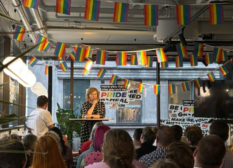 Kuvan keskellä Kati Laitinen seisoo lavalla. Hän nojaa pöytään ja puhuu mikrofoniin. Lavan yläpuolelle kattoon on ripustettu Pride-lippuja.