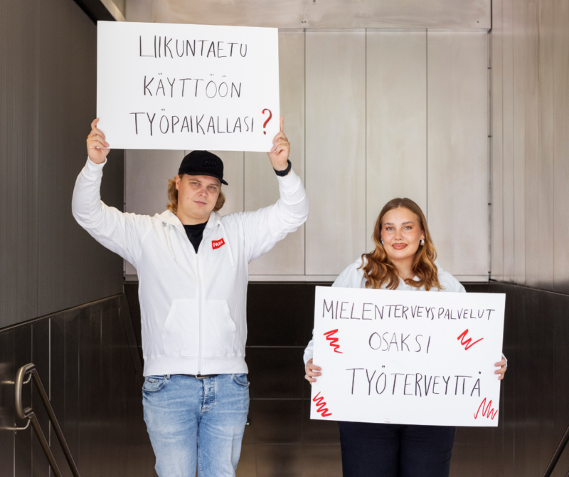 Kuvassa kaksi PAMin ohjausryhmän nuorta pitävät kahta kampanjakylttiä käsissään. Niissä lukee: "Liikuntaetu käyttöön työpaikallasi?" ja "Mielenterveyspalvelut osaksi työterveyttä".