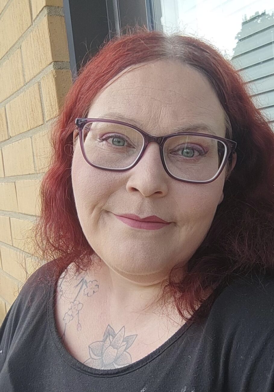 Lähikuva punahiuksisesta naisesta, jolla on silmälasit ja harmaa paita.