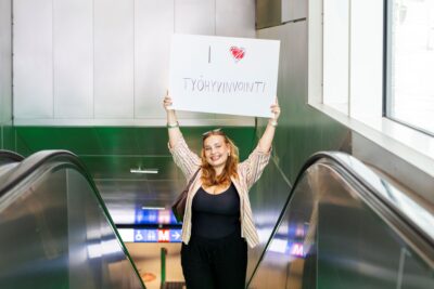 Nuori nainen seisoo liukuportaissa ja pitää kylttiä, jossa lukee I