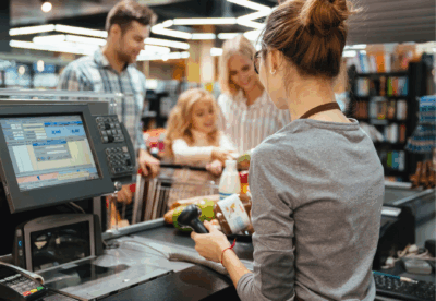 Päivitetystä oppaasta poimittu kuvituskuva, jossa työntekijä piippaa tuotteita kaupan kassalla. Taustalla perhe.