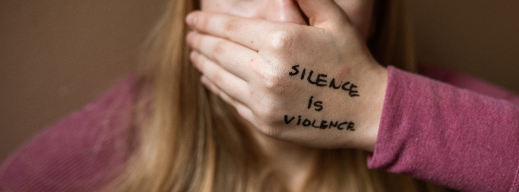 Nainen peittää suunsa kädellään. Kädessä lukee "silence is violence".