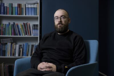 Fred Miller, Chair of PAM’s Impact Forum, sitting with a serious expression, a blurred bookshelf in the background.