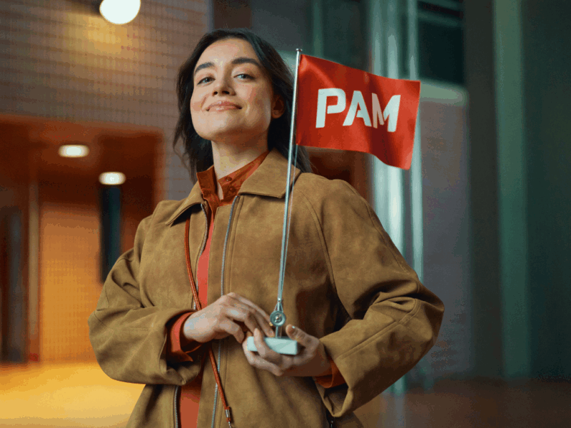 Nuori aikuinen seisoo itsevarmana kaupunkimiljöössä ja pitelee pientä punaista PAM-lippua. Hänellä on ruskea takki ja lämmin ilme, ja taustalla näkyy moderni rakennus iltavalaistuksessa.