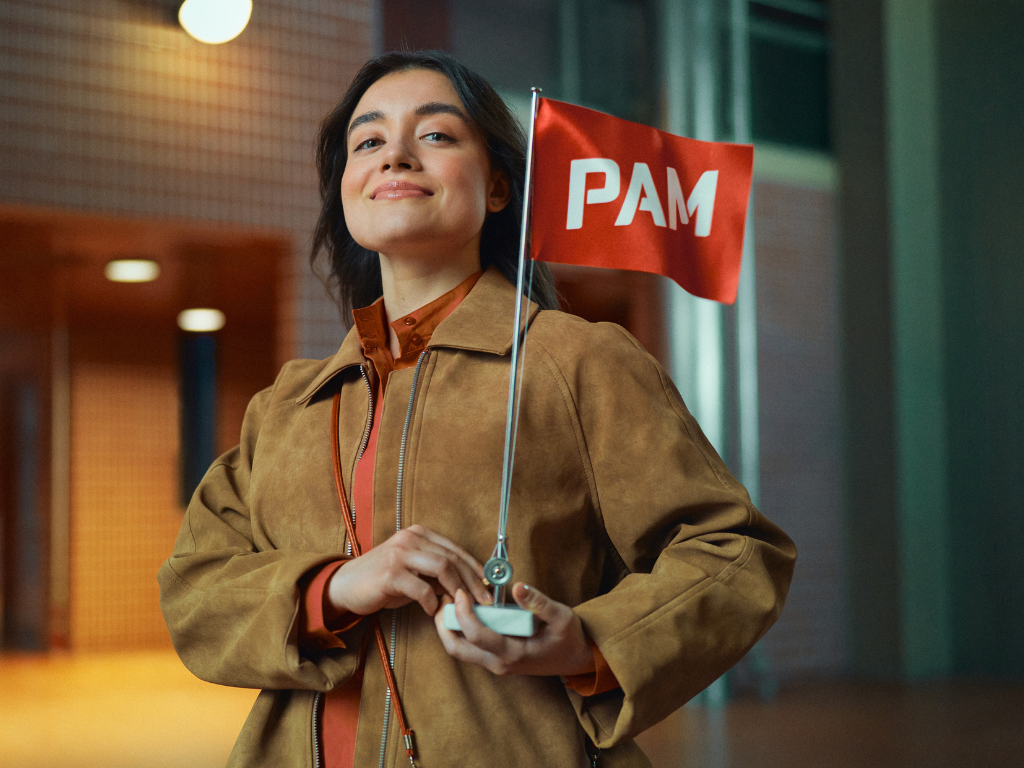 Nuori aikuinen seisoo itsevarmana kaupunkimiljöössä ja pitelee pientä punaista PAM-lippua. Hänellä on ruskea takki ja lämmin ilme, ja taustalla näkyy moderni rakennus iltavalaistuksessa.