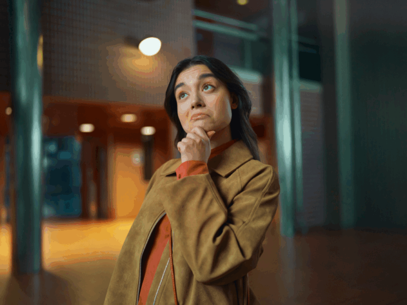 Nuori aikuinen seisoo sisätilassa ja katsoo mietteliäänä ylöspäin käsi leukaa vasten. Hänellä on ruskea takki, ja taustalla näkyy moderni rakennus lämpimässä valaistuksessa.