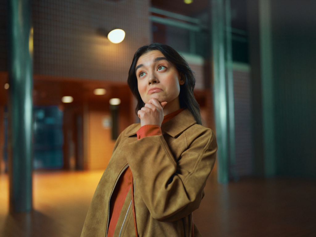 Nuori aikuinen seisoo sisätilassa ja katsoo mietteliäänä ylöspäin käsi leukaa vasten. Hänellä on ruskea takki, ja taustalla näkyy moderni rakennus lämpimässä valaistuksessa.