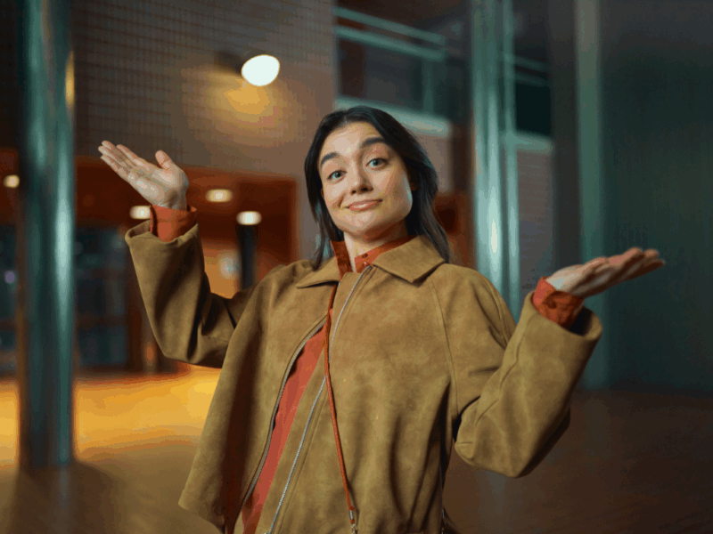 Nuori aikuinen seisoo sisätilassa kädet levällään ja hymyilee ilmeellä, joka viestii "mikäpä ettei" -ilmettä. Hänellä on ruskea takki ja taustalla näkyy moderni rakennus lämpimässä valaistuksessa.