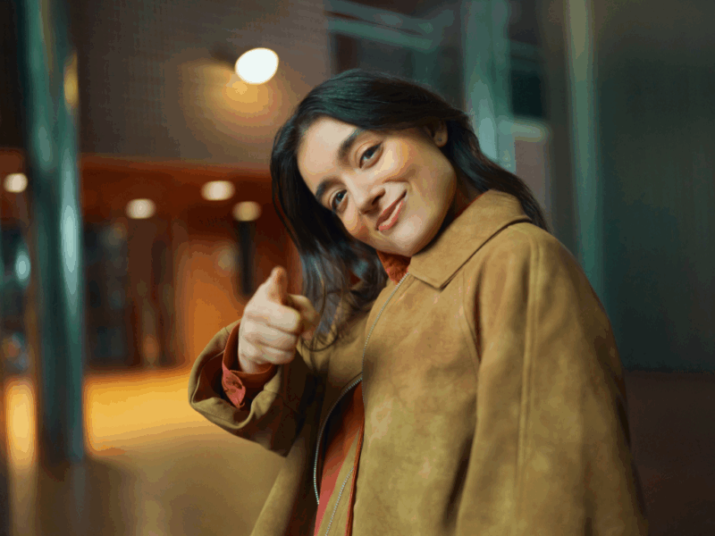 Nuori aikuinen hymyilee ja osoittaa kameran suuntaan etusormellaan. Hänellä on ruskea takki ja taustalla näkyy moderni rakennus pehmeässä iltavalaistuksessa.