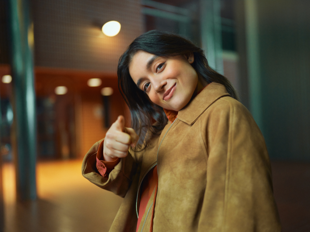 Nuori aikuinen hymyilee ja osoittaa kameran suuntaan etusormellaan. Hänellä on ruskea takki ja taustalla näkyy moderni rakennus pehmeässä iltavalaistuksessa.