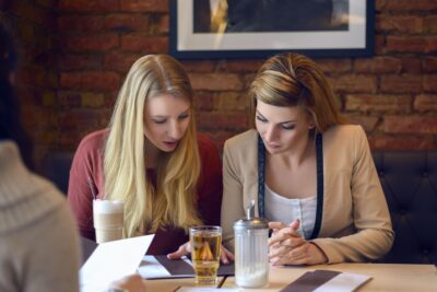 Kaksi vaaleatukkaista naista istuvat pöydän ääressä ja tarkastelevat papereita.