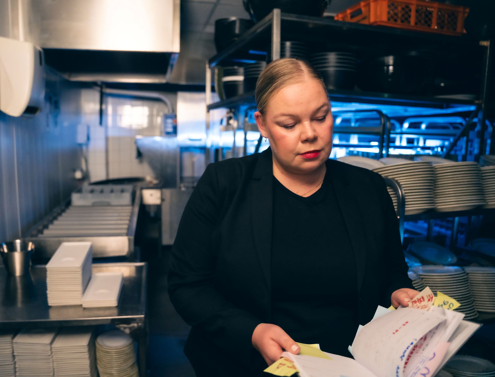 Ravintolatyöntekijä seisoo keittiön työtilassa ja tarkistaa papereita katse keskittyneenä.