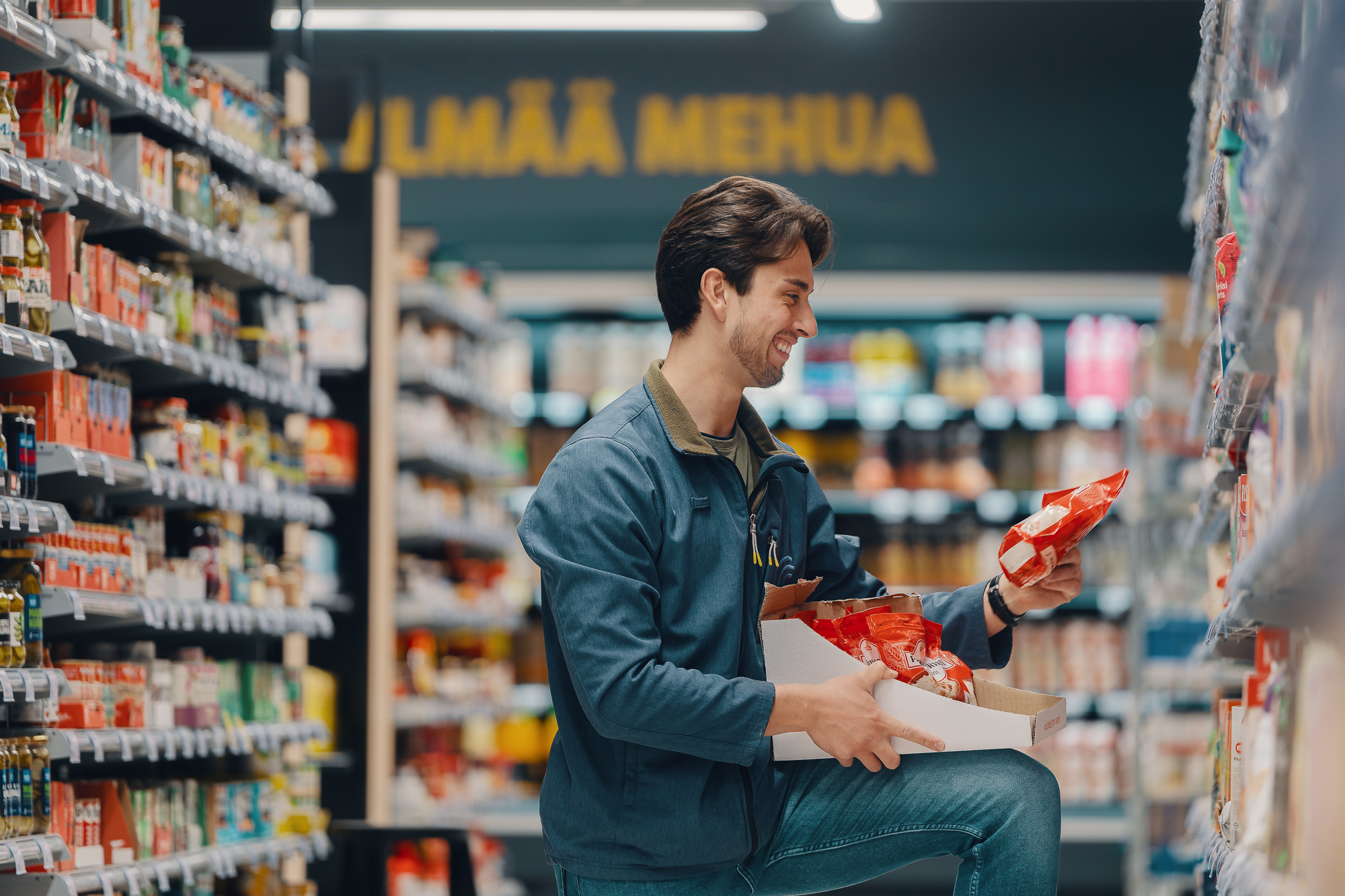 Kaupan työntekijä polvistuneena hyllyttää punaisia elintarvikepakkauksia ruokakaupan käytävällä, pitää laatikkoa sylissään ja tarkistaa tuotetta hymyillen.