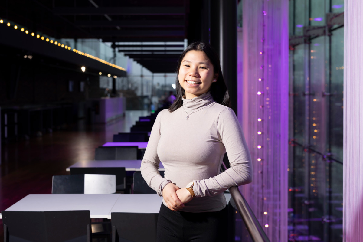 Nuori nainen hymyilee ja seisoo kädet edessä ikkunan vieressä modernissa ravintolatyyppisessä tilassa, taustalla pöytiä, tuoleja ja violettia tunnelmavalaistusta.