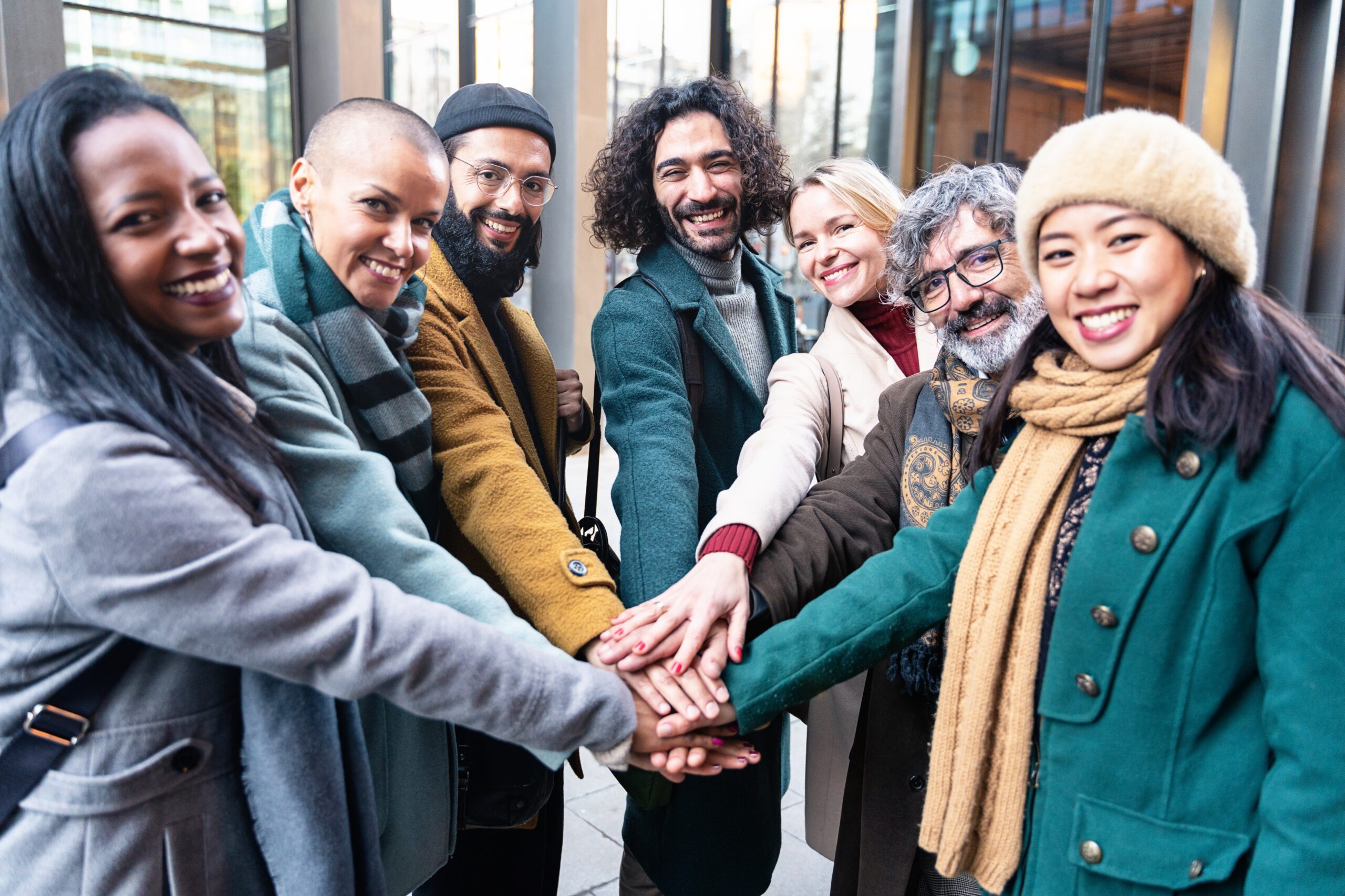 Seven diverse people of different ages and backgrounds stand in a circle outside a building, smiling and stacking their hands together in the center to show solidarity and teamwork.