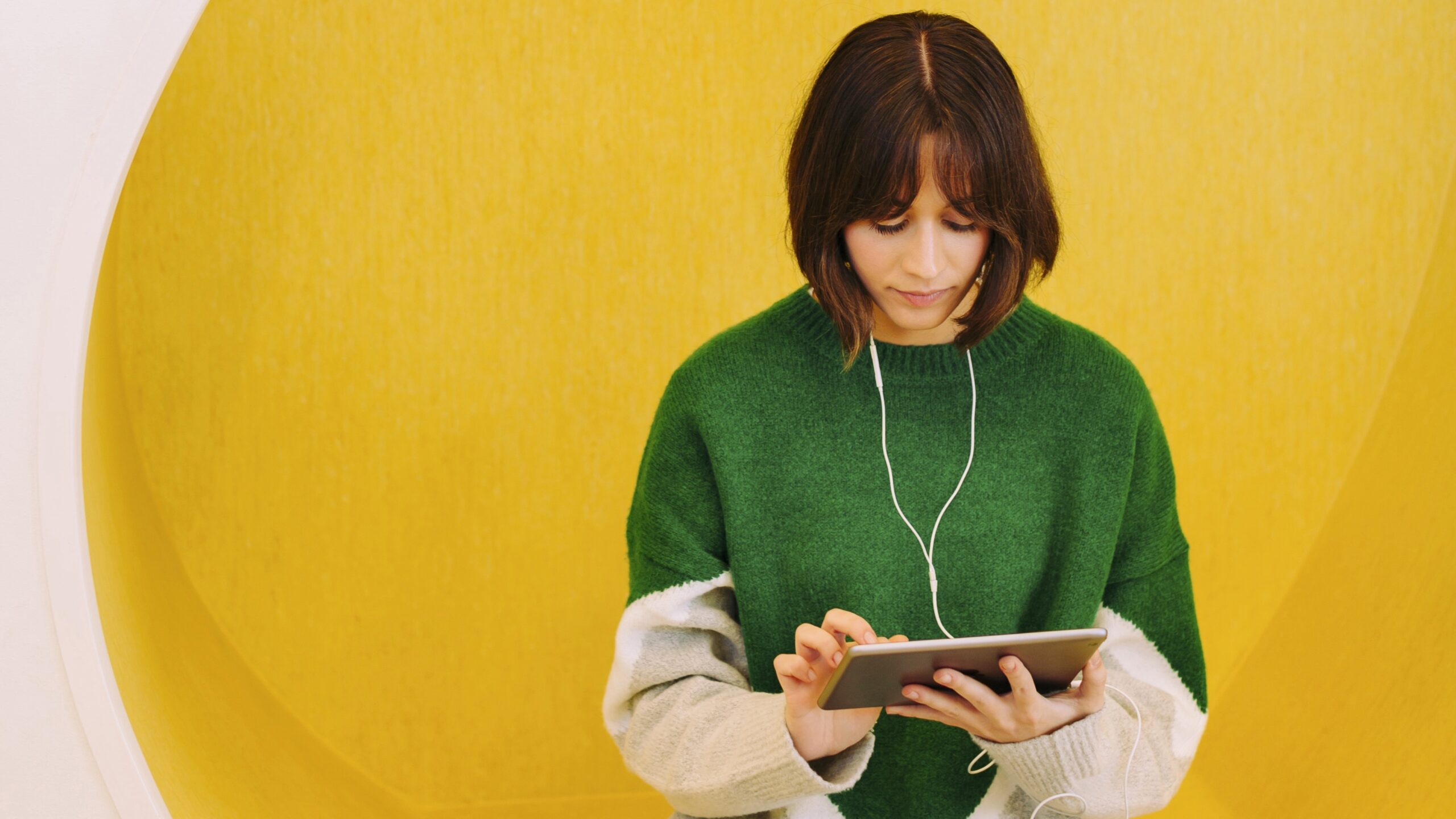 Nainen vihreässä neuleessa istuu keltaisessa pyöreässä aukossa, kuuntelee kuulokkeilla ja käyttää tablettia.