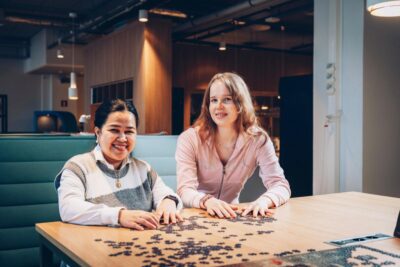 Kaksi naista istuu modernissa työtilassa pöydän ääressä ja kokoaa palapeliä, hymyillen - kuva symboloi ammattiliiton ja työttömyyskassan erojen selvittämistä.