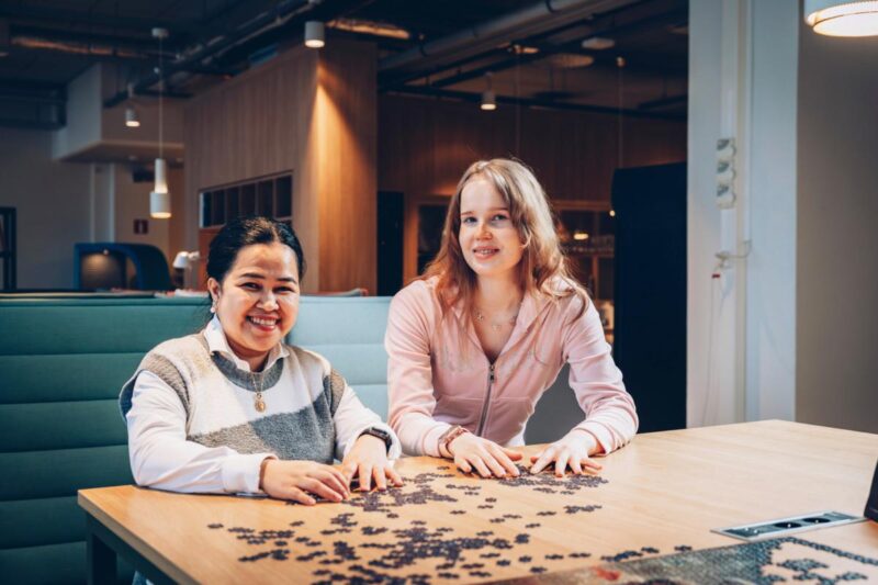 Kaksi naista istuu modernissa työtilassa pöydän ääressä ja kokoaa palapeliä, hymyillen - kuva symboloi ammattiliiton ja työttömyyskassan erojen selvittämistä.