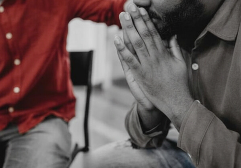 Close-up of two seated people in an office: a person in the foreground leans forward with hands clasped near their mouth in a tense, worried posture, while the other person beside them rests a reassuring hand on their shoulder, conveying concern and support.