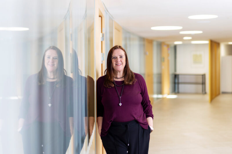 Carol Scheffer nojaa lasiseinään modernissa ja valoisassa käytävässä, hymyilee ja pitää käsiä taskuissa, hänen heijastuksensa näkyy lasipinnassa.