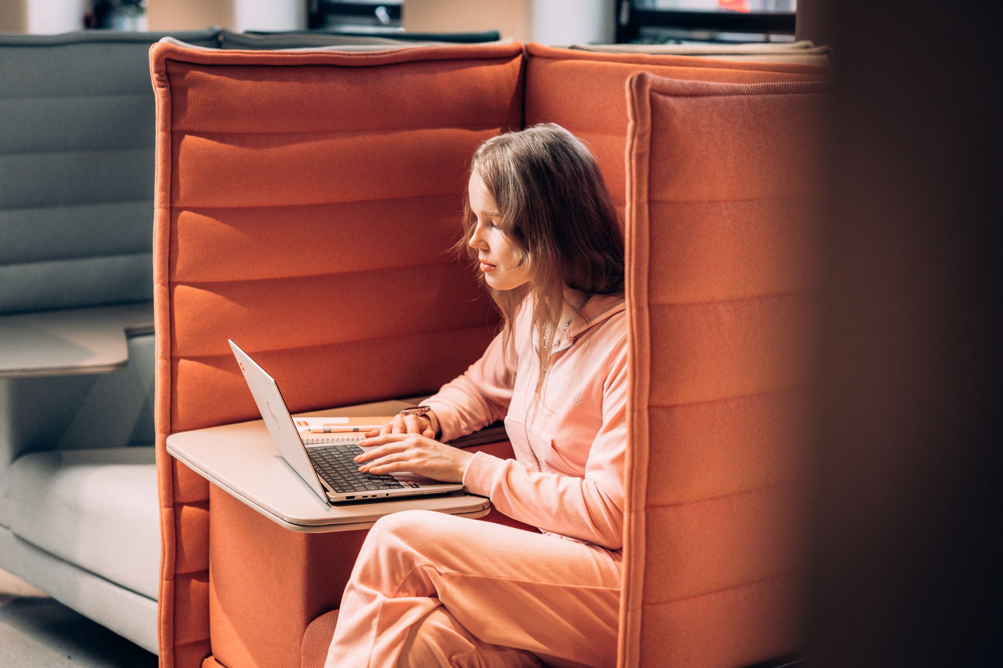 Henkilö istuu oranssissa äänieristetyssä sohvanurkkauksessa keskittyneenä kannettavan tietokoneen ääressä.