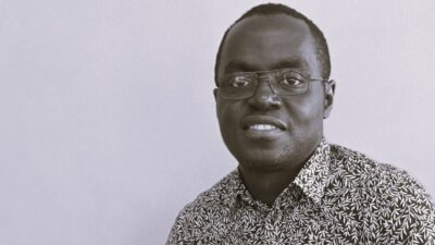 Head-and-shoulders portrait of Germain Ishimwe, a smiling man with short hair and glasses wearing a patterned shirt, set against a plain light background.