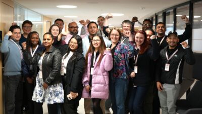Group photo with about 20 PAM Network Leaders in an office corridor, smiling and some raising fists, wearing lanyards and casual clothes.