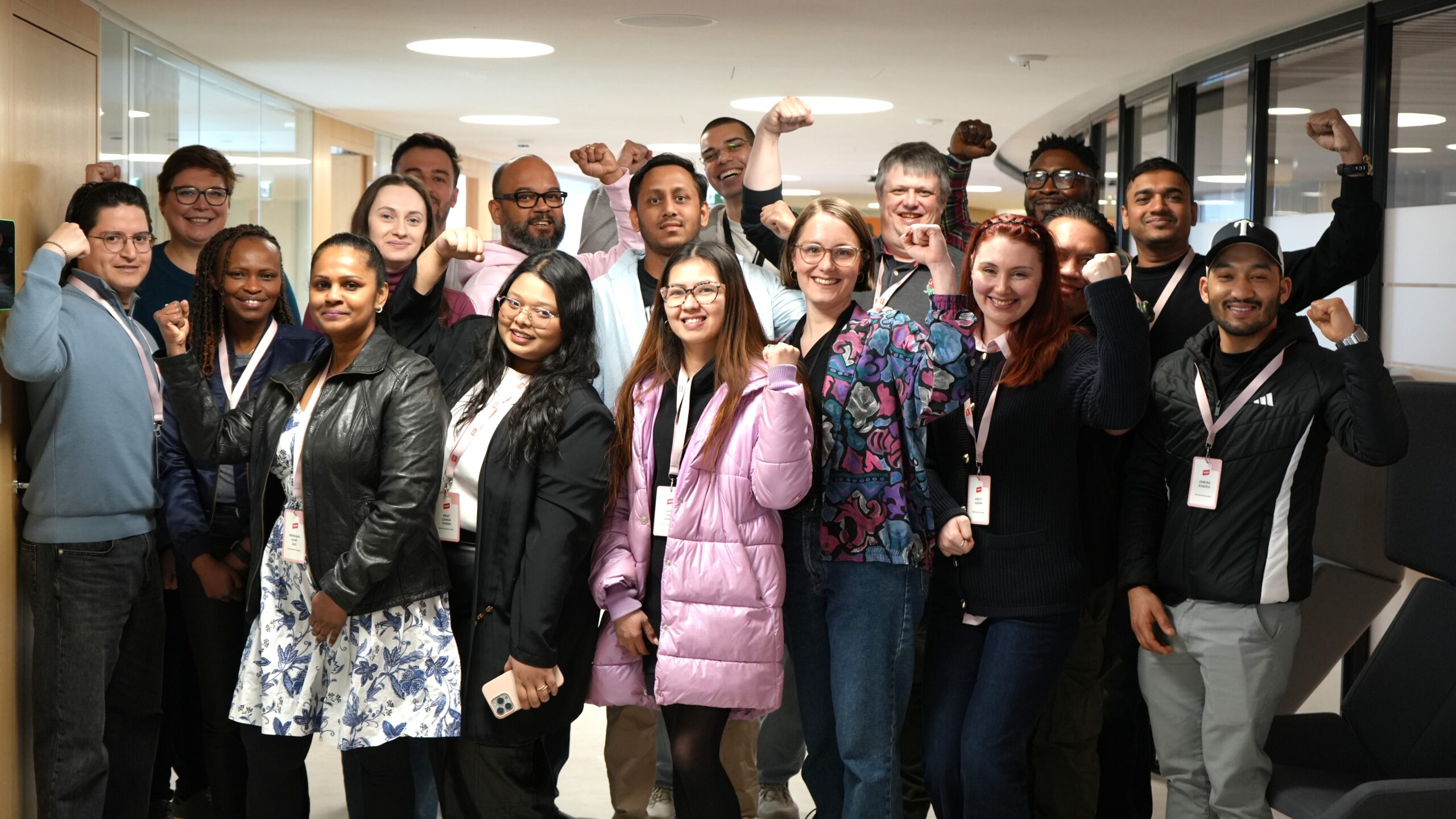 Group photo with about 20 PAM Network Leaders in an office corridor, smiling and some raising fists, wearing lanyards and casual clothes.