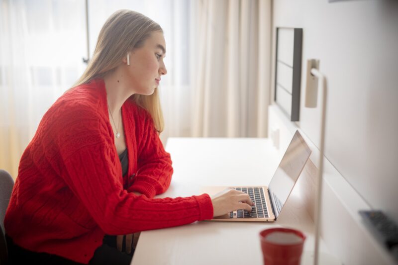Ung kvinna i röd tröja sitter vid skrivbord och arbetar på sin bärbara dator, en röd mugg i förgrunden och mjukt dagsljus från fönstret i bakgrunden.