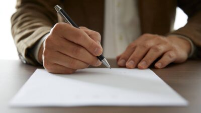 Kuvassa näkyy henkilön kaksi kättä nojaamassa pöydällä olevaa paperia vasten. Toisessa kädessä on kynä valmiina allekirjoittamaan sopimuksen.