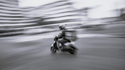Skootterilähetti kypärä päässä ja takalaukku selässä ajaa kovaa kaupunkikadulla; kuva on liike-epäterävä ja korostaa nopeutta ja työssä olevaa riskiä.
