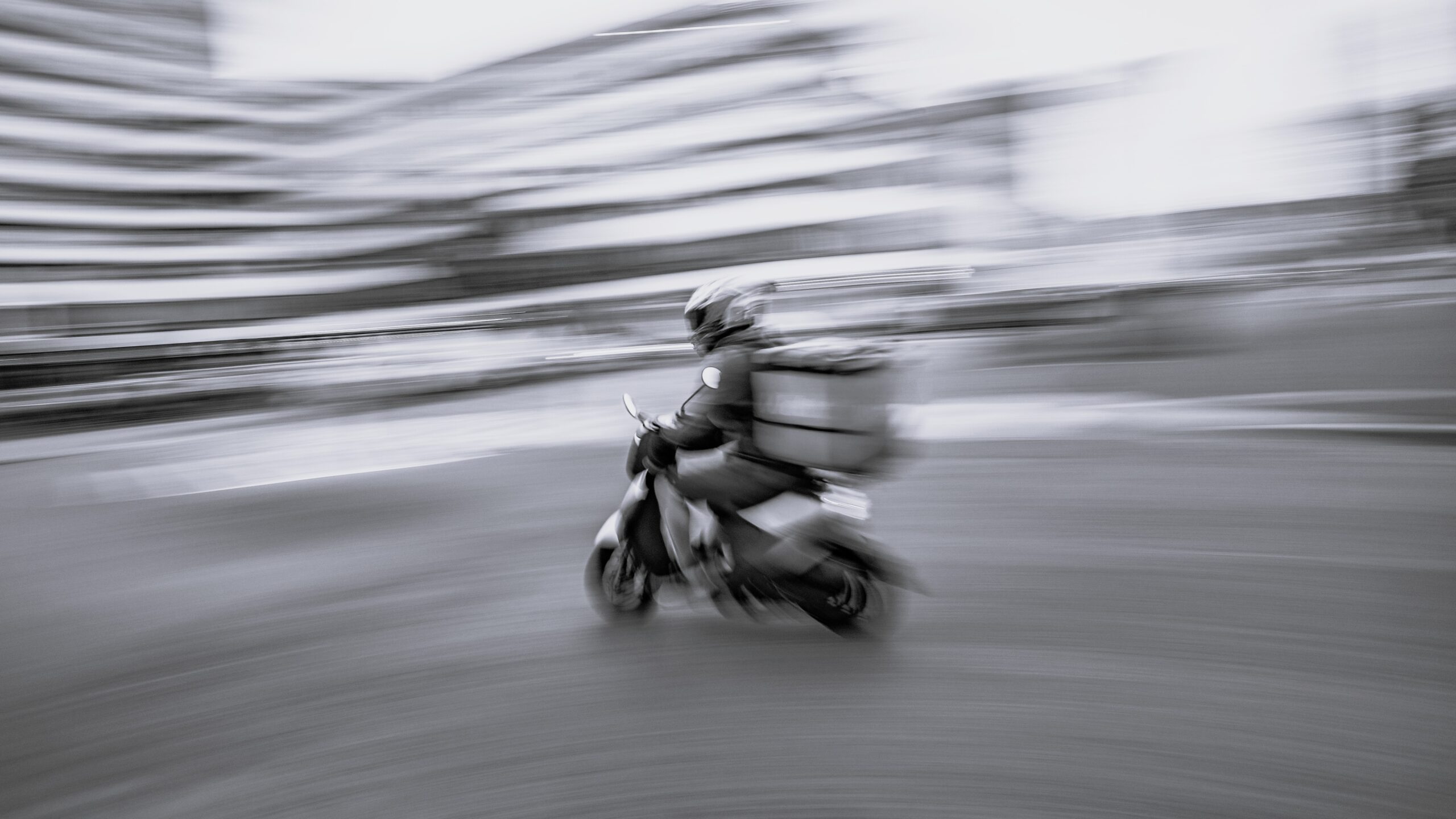 Skootterilähetti kypärä päässä ja takalaukku selässä ajaa kovaa kaupunkikadulla; kuva on liike-epäterävä ja korostaa nopeutta ja työssä olevaa riskiä.