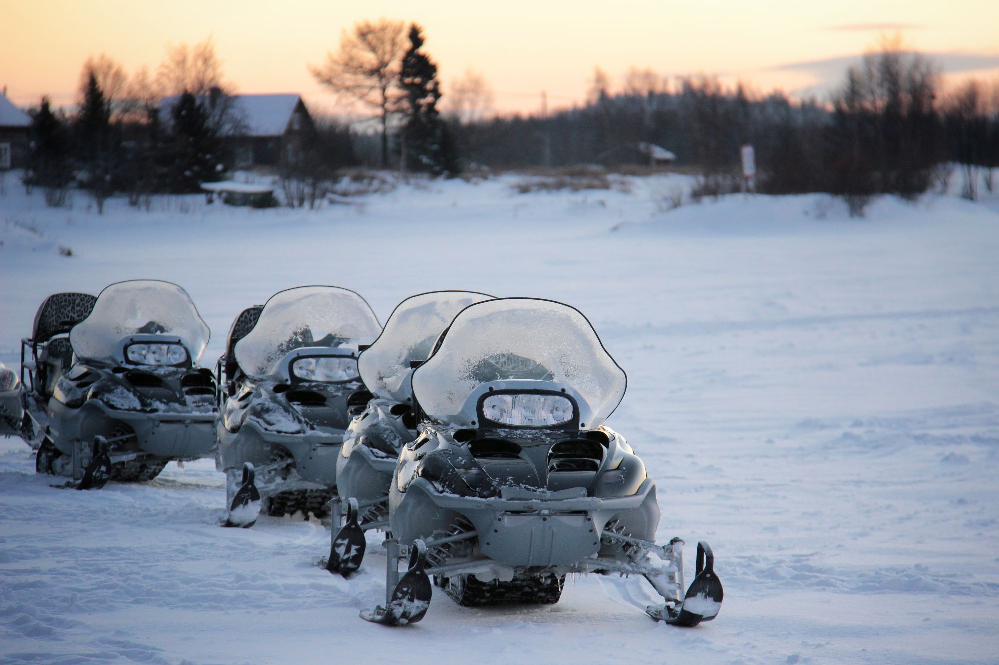 Kolme lumipeitteistä moottorikelkkaa rivissä talvisella kentällä, etualalla hanki ja taustalla hämärtyvä mökki- ja metsänäkymä