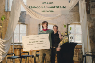 Kaksi henkilöä kaulakkain. Toisella kukat kädessä ja toisella iso plakaatti, jossa kerrotaan myönnetystä tunnustuksesta.