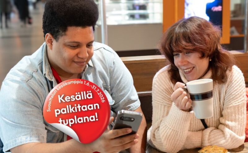 Kaksi hymyilevää aikuista istuu kahvilapöydässä ja katsoo yhdessä puhelinta, toinen pitelee mukia, kuvan päällä punainen merkki tekstillä "Kesällä palkitaan tuplana!"—yhteisöllinen ja kannustava tunnelma.