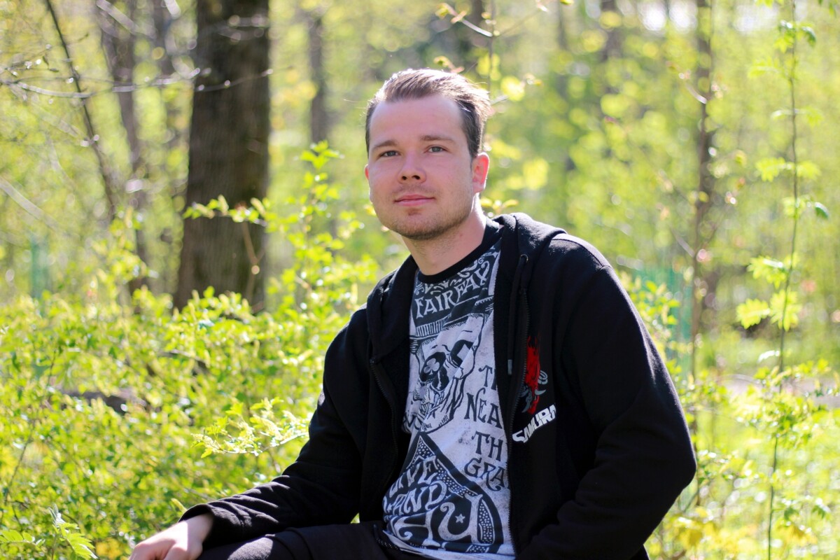 Nuori mies istuu keväisessä metsäympäristössä aurinkoisen värikkään aluskasvillisuuden keskellä, katsoo kameraan rauhallinen ja luottavainen ilme, päällään huppari ja kuvioitu t-paita.