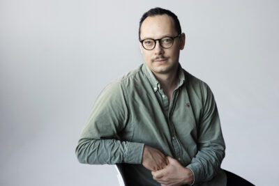 Paavo Teittinen istuu tuolilla ja katsoo suoraan kameraan, kädet edessä yhteenpujotettuina; hänellä on vihreä kauluspaita, pyöreät silmälasit ja vaalea neutraali tausta.