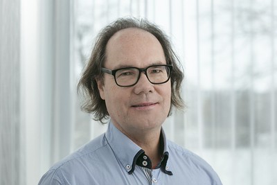 Rintakuva hymyilevästä Juha Antilasta sinisessä kauluspaidassa, vaalea ja sumennettu ikkunatausta.