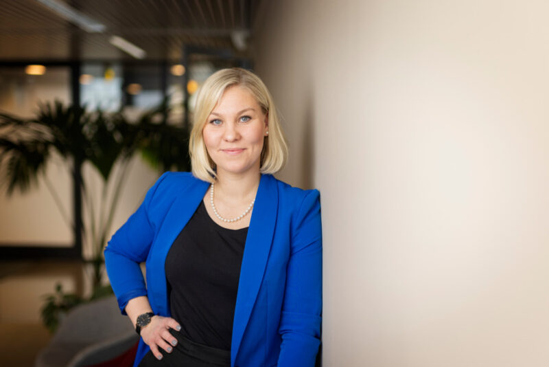 Hymyilevä, vaaleahiuksinen nainen sinisessä bleiserissä nojaa toimiston seinään käsi lantiolla, taustalla kasveja ja toimistotilaa.