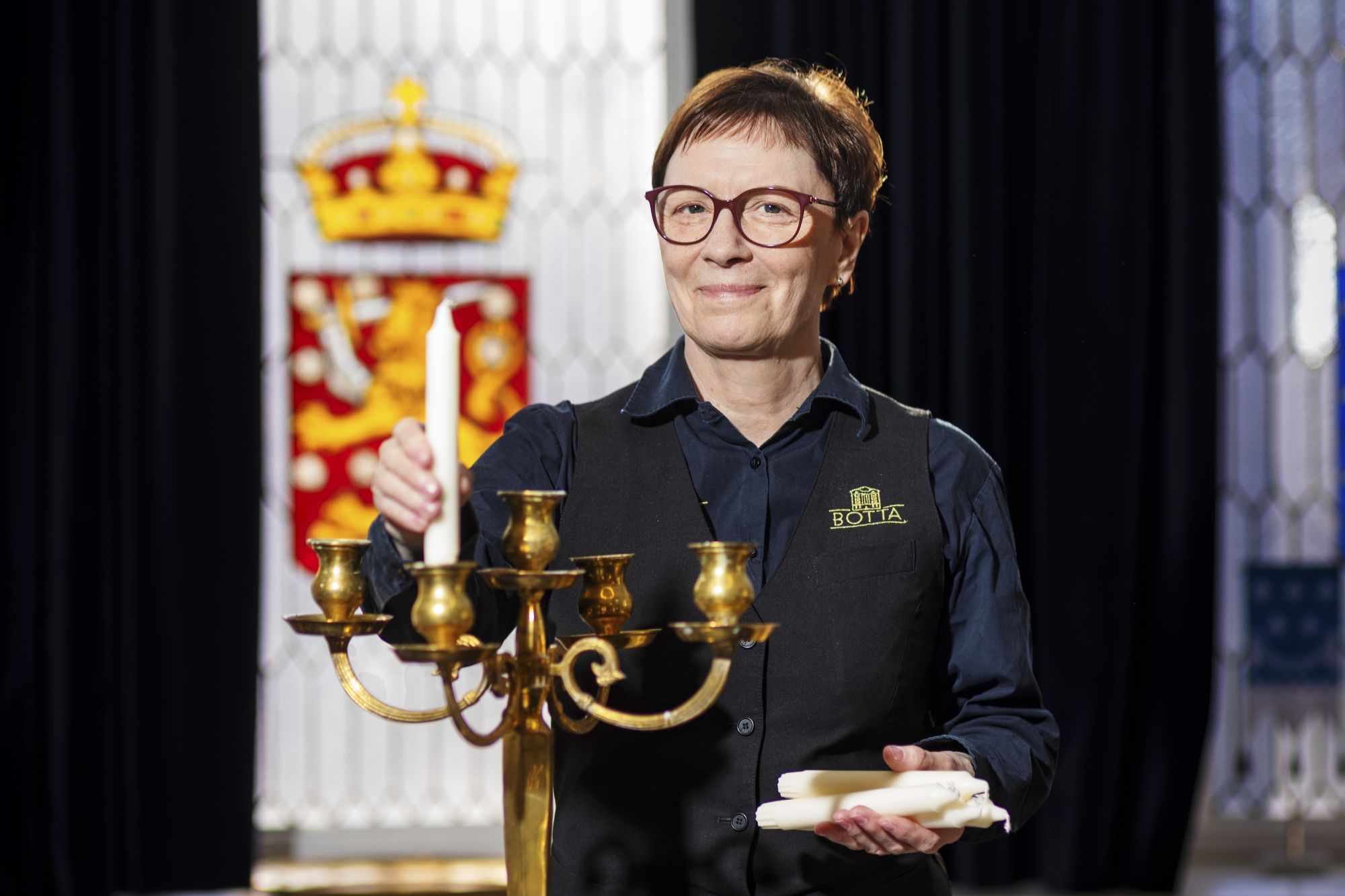Palvelualan työntekijä tumma työasu päällään hymyilee ja puhdistaa liinalla messinkistä viisihaaraista kynttilänjalkaa, taustalla lasimaalauksellinen ikkuna jossa näkyy Suomen vaakuna