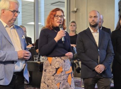Tapahtumatilassa seisova Annika Rönni-Sällinen puhuu mikrofoniin, hänen sivullaan kaksi miestä kuuntelee ja taustalla näkyy ihmisiä sekä tarjoiluja.