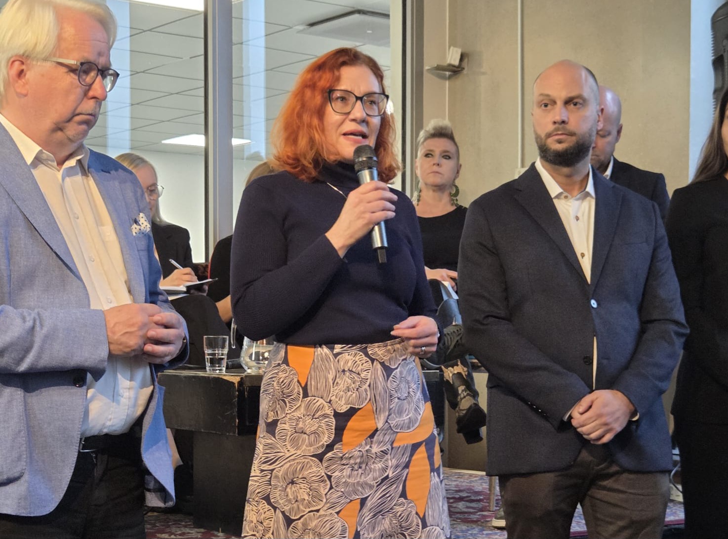 Tapahtumatilassa seisova Annika Rönni-Sällinen puhuu mikrofoniin, hänen sivullaan kaksi miestä kuuntelee ja taustalla näkyy ihmisiä sekä tarjoiluja.